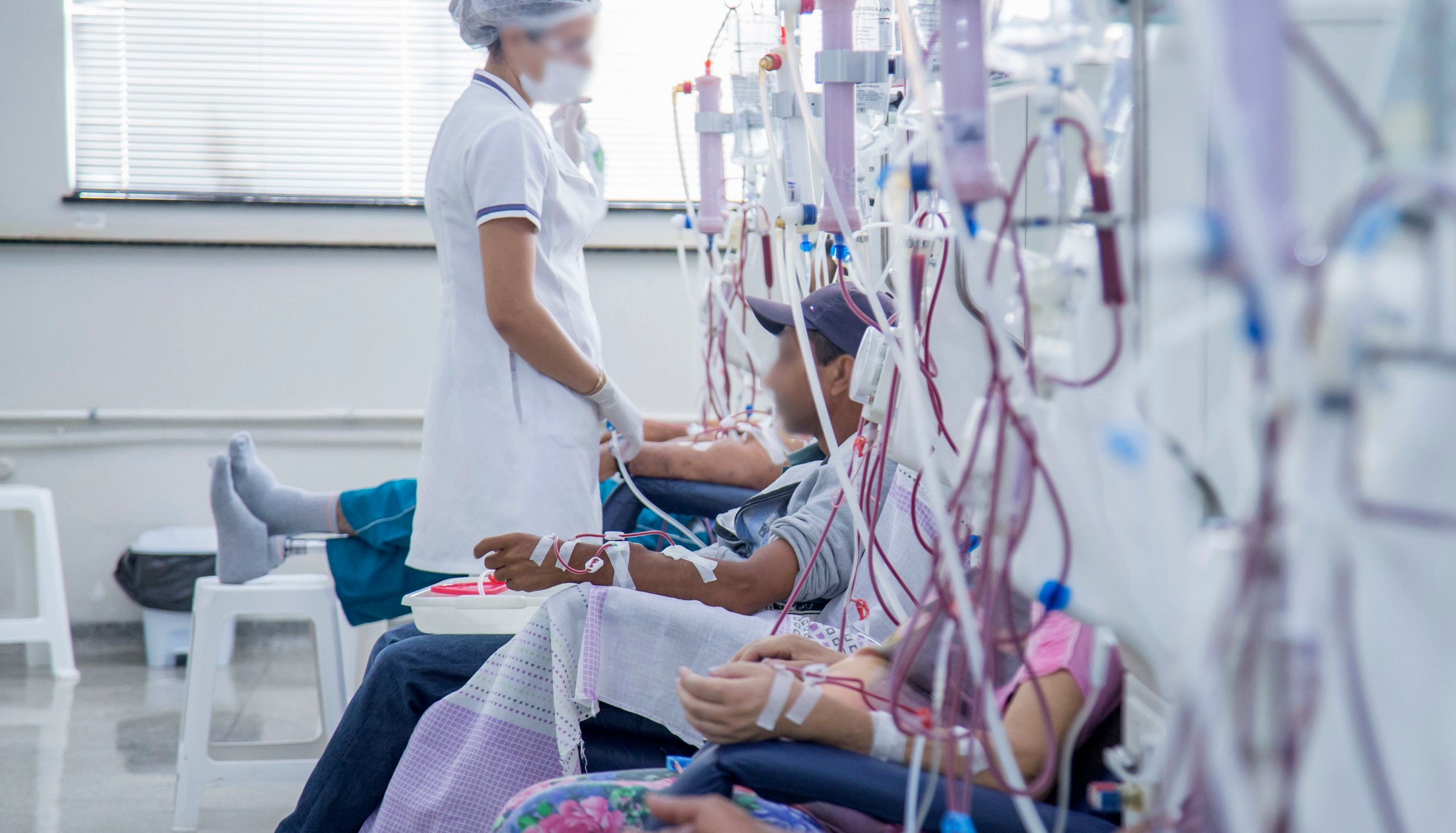 Patients seated in a row receiving dialysis, connected by tubing to machines while a nurse attends them